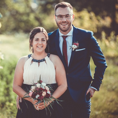 Frisch verheiratetes Brautpaar posiert lächelnd im Freien bei weichem Gegenlicht mit Brautstrauß.