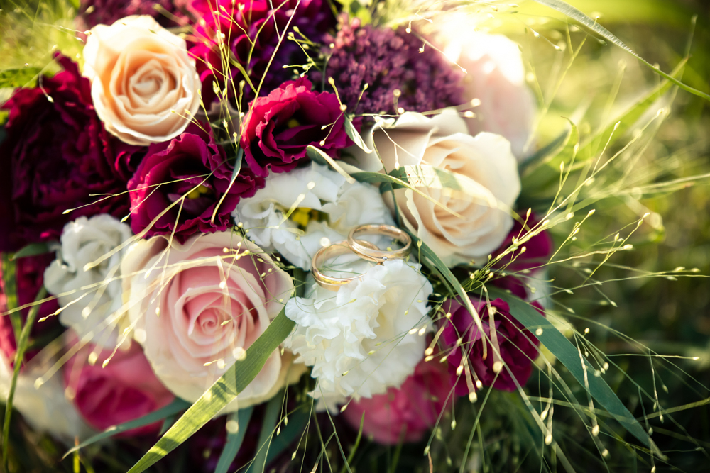 Zwei Eheringe liegen auf einem Hochzeitsstrauß aus Rosen und Nelken im Gegenlicht.