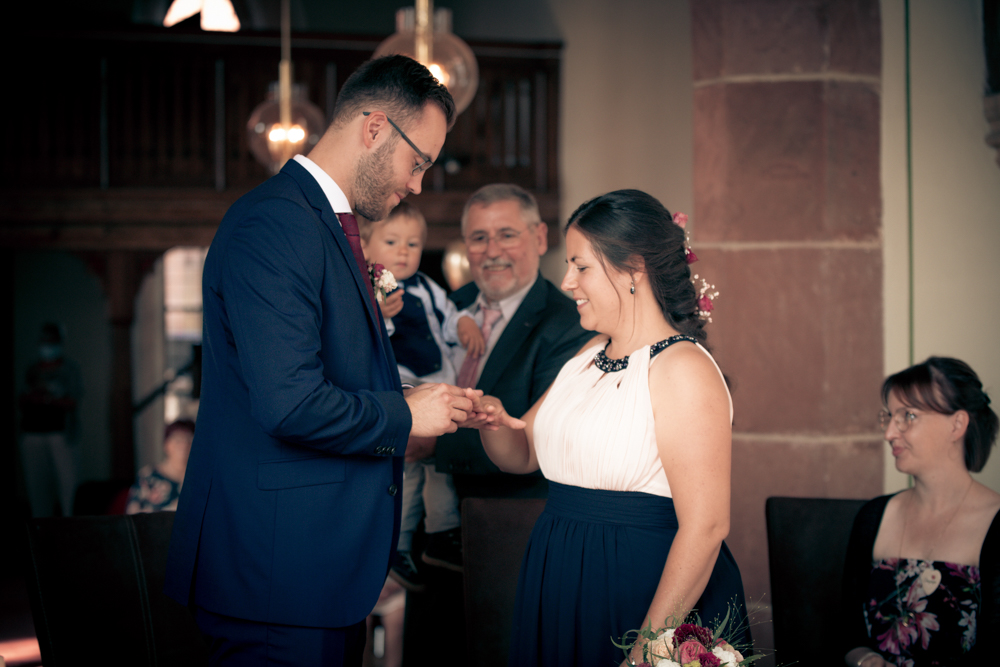 Bräutigam steckt der Braut feierlich den Ring an – ihr Kind auf dem Arm beobachtet die Zeremonie.