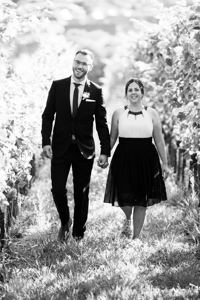 Ein Brautpaar spaziert Hand in Hand durch die Weinberge in der Pfalz und lacht gemeinsam.
