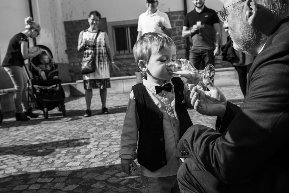 Ein kleiner Junge im Anzug trinkt aus einem Sektglas, das ihm ein Erwachsener reicht. Schwarzweißaufnahme mit Gästen im Hintergrund.