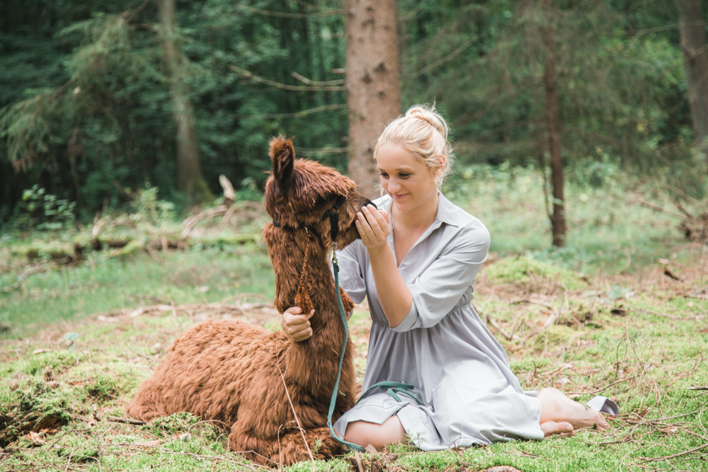 Portraitfotografie mit einer jungen Frau, die neben einem Lama im Wald sitzt und es liebevoll streichelt.