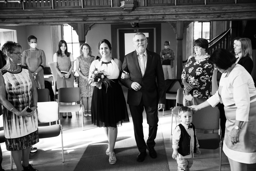 Schwarz-Weiß-Fotografie einer Frau, die von einem älteren Mann zum Altar geführt wird, während Gäste in einer Kirche zusehen.